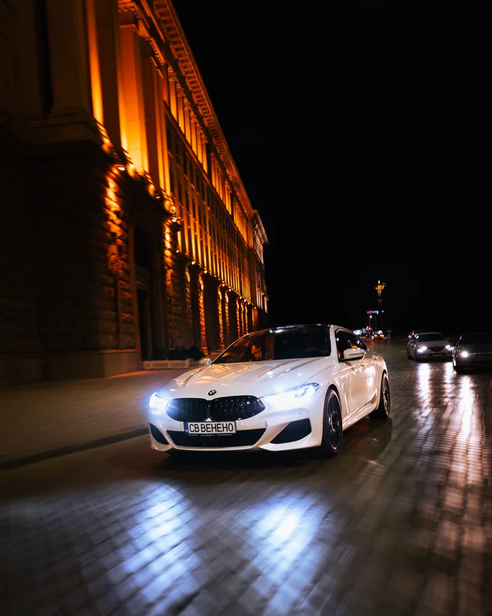 Бяло BMW нощем на улица с калдъръм пред осветена сграда
